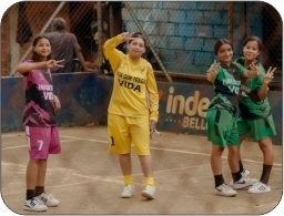 Four girls wearing sport uniforms in a fenced in sports area smiling at the camera